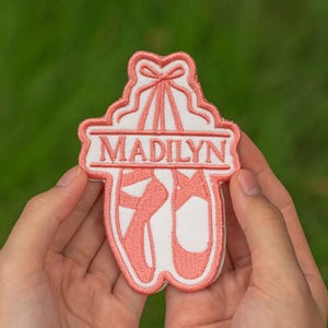 May include: Embroidered patch featuring ballet shoes and the name "MADILYN" in a pink and white design. The patch has a bow at the top and is held in a person's hands against a blurred green background. The patch is approximately 3 inches in height.