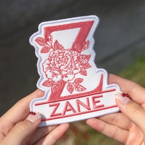 May include: Embroidered patch featuring the name "ZANE" in red, with a stylized "Z" and a floral rose design. The patch has a white border and is held in a person's hands.