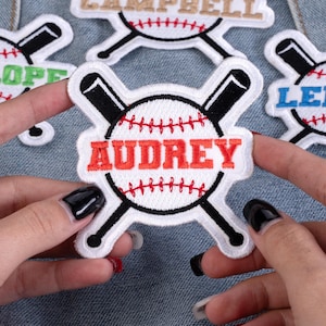 May include: Embroidered baseball patch with the name "AUDREY" in red letters. The patch features a white baseball design with red stitching and crossed black bats. Other patches with names are visible in the background.