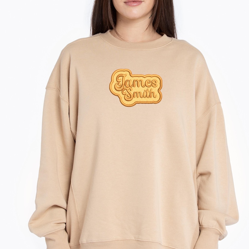 May include: A beige sweatshirt with a retro-style graphic of the name "James Smith" in a yellow and orange color scheme.