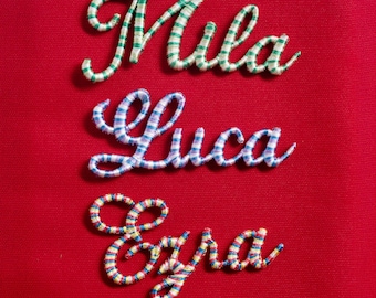 May include: Embroidered names Mila, Luca, and Ezra in script font on a red background. The names are stitched with colorful thread: green and white, blue and white, and a mix of red, yellow, and blue. The names are likely for personalization.