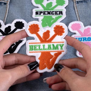 May include: Embroidered patches featuring a cheerleader design in orange, green, black, and pink. Each patch has a name in a rectangular banner. The patch held in the center reads "Bellamy" in green letters.