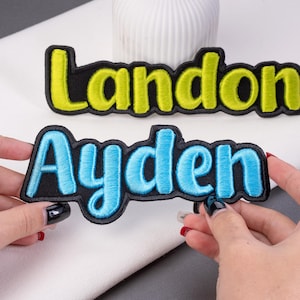 May include: Embroidered name patches with black borders. One patch reads "Landon" in yellow letters, and the other reads "Ayden" in blue letters. The patches are on a white surface.