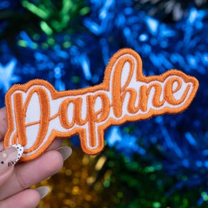 May include: An embroidered patch with the name "Daphne" in orange script lettering on a white background. The patch has an orange border and is held against a blurred background of blue and green.