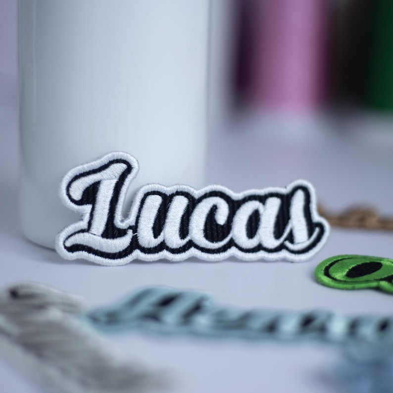 May include: White embroidered patch with the name "Lucas" in black outlined lettering.