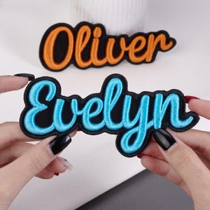 May include: Two embroidered name patches. One patch reads "Oliver" in orange thread with a black border. The other patch reads "Evelyn" in blue thread with a black border. Both patches have a cursive font.