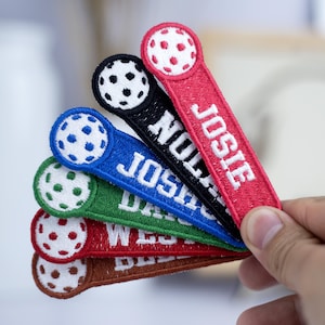 May include: Seven colorful embroidered patches with a white ball and black stitching. Each patch has a different name: Josie, Nolan, Josiah, Dylan, Wesley, and two others.