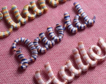 May include: Close-up of embroidered names on a pink fabric. The names are stitched in a cursive style with colorful thread. The thread colors include blue, orange, and white. The names are "David" and "Hello".