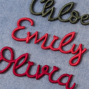 May include: Close-up of embroidered names in cursive script. The names "Chloe" in olive green, "Emily" and "Olivia" in red thread are displayed on a light blue denim background. The embroidery has a raised, textured appearance.