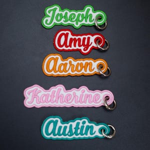 May include: Five embroidered fabric keychains with white borders and silver metal rings. Each keychain features a different name in cursive script: Joseph in green, Amy in red, Aaron in orange, Katherine in light pink, and Austin in teal.