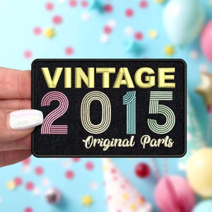 May include: A black rectangular patch with the words "VINTAGE 2015 Original Parts" in yellow, pink, teal, and white. The patch is held against a light blue background with colorful confetti and balloons.