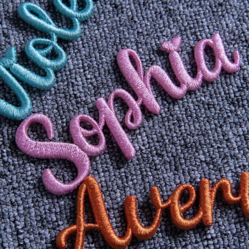 May include: Close-up of embroidered names in cursive script. The name "Sophia" is in pink, "Avery" is in orange, and another name is in teal. The names are stitched on a textured gray fabric.