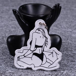 May include: White embroidered keychain featuring a black line drawing of a person in lingerie. The keychain has a silver metal ring for attachment. The design is on a white background with a black border.
