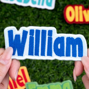 May include: A blue embroidered patch with the name "William" in bold, capital letters. The patch has a light blue border and is held by a person's hands. Other name patches are visible in the background.