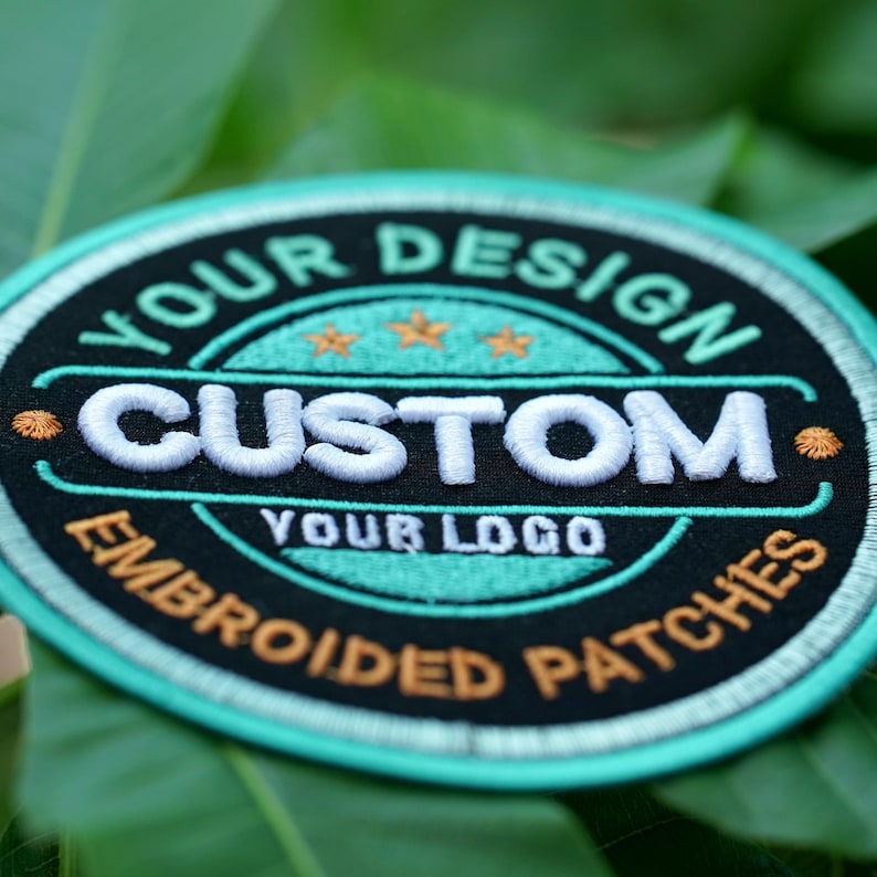 Pu&ograve; includere: Toppa ricamata circolare con la scritta "YOUR DESIGN CUSTOM YOUR LOGO EMBROIDERED PATCHES". La toppa ha uno sfondo nero con accenti bianchi, verde acqua e arancioni. La toppa &egrave; su uno sfondo di foglie verdi.