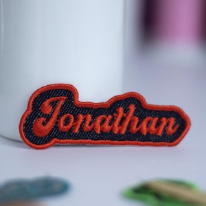 May include: A custom embroidered patch with the name "Jonathan" in red script lettering. The name is stitched on a navy blue background with a red border. The patch is a personalized item.