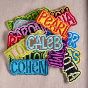May include: A collection of embroidered name patches in various colors and fonts, including names such as Pearl, Caleb, and Cohen. Each patch features a thick, stitched border and bold lettering, layered together in a dense, overlapping arrangement.