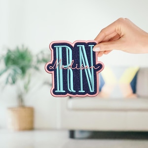 May include: Embroidered patch featuring the letters "RN" in a light blue color, with the name "Madison" in a cursive font. The patch has a navy blue background and a pink border. The patch is held by a hand.