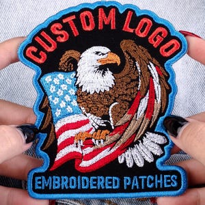 May include: Embroidered patch featuring a detailed bald eagle clutching an American flag. The patch has the text "CUSTOM LOGO" in red and "EMBROIDERED PATCHES" in blue. The eagle is brown, white, and orange, with a blue border.