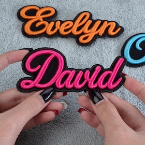 May include: Embroidered name patches in orange, pink, and blue. The pink patch, held by hands, reads "David" in a cursive font with a black border. An orange patch above reads "Evelyn".