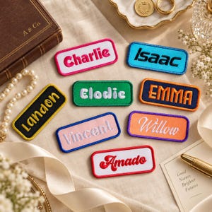 May include: A collection of rectangular embroidered name patches featuring various colors and fonts. The patches display the names Charlie, Isaac, Landon, Elodie, Emma, Vincent, Willow, and Amado. Each patch has a distinct border color and text style.