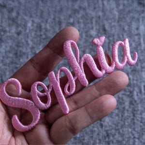 May include: A pink embroidered patch spelling "Sophia" in a cursive font. The patch is held in a hand against a gray background. The letters are in a delicate, flowing script, and the thread has a slight sheen.