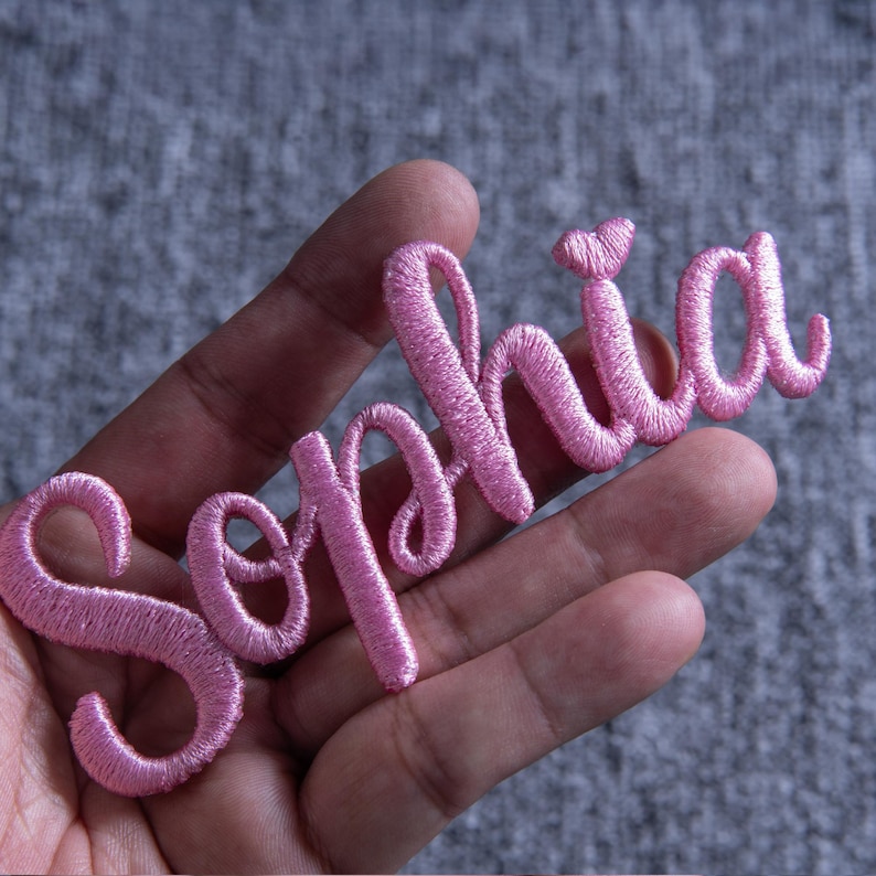 May include: A pink embroidered patch spelling "Sophia" in a cursive font. The patch is held in a hand against a gray background. The letters are in a delicate, flowing script, and the thread has a slight sheen.