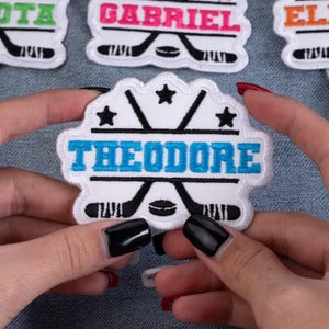 May include: Embroidered patch with the name "THEODORE" in blue, outlined in white, on a white background. The patch features hockey sticks, a puck, and stars. The patch is held by a person.