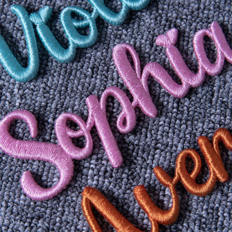 May include: Close-up of embroidered names in various colors. The name "Sophia" is in pink, "Aver" is in orange, and a portion of a teal name is visible. The embroidery is on a textured gray fabric, showcasing the detailed stitching.