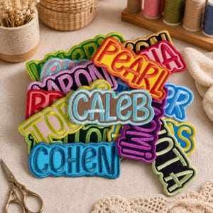 May include: A collection of embroidered name patches in various colors and fonts, including names like Pearl, Caleb, and Cohen. These fabric patches feature stitched borders and bold lettering, layered together in a pile on a textured fabric surface.