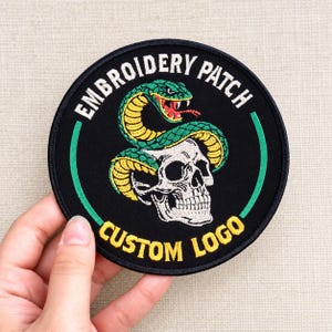 May include: A circular black embroidered patch featuring a white skull wrapped in a green and yellow snake. The text "EMBROIDERY PATCH" is arched in white at the top, and "CUSTOM LOGO" is arched in yellow at the bottom, bordered by green lines.