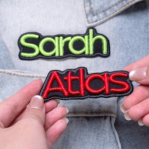 May include: Two embroidered name patches with black backgrounds and raised borders. One patch features the name Sarah in bright lime green thread, while the other patch displays the name Atlas in vibrant red thread, both in a rounded font.