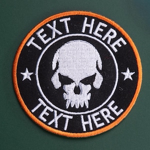 May include: A circular embroidered patch featuring a white skull graphic in the center, surrounded by the white text "TEXT HERE" at the top and bottom. The design includes two white stars on the sides, all set against a black background with a bright orange border.