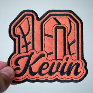 May include: Orange and black basketball-themed iron-on patch with the number "10" and the name "Kevin" in a black script font.