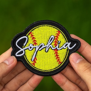 May include: Embroidered patch featuring a yellow softball design with red stitching and the name "Sophia" in white script. The patch has a black border and is held against a green background.