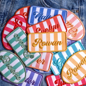 May include: A collection of rectangular and oval embroidered patches featuring white vertical stripes alternating with various colors including blue, red, orange, green, and purple. Each patch displays a unique name embroidered in cursive script.