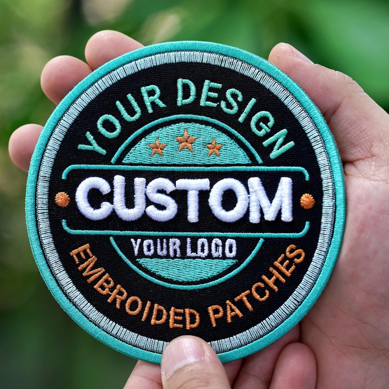 Pu&ograve; includere: Toppa rotonda ricamata con la scritta "YOUR DESIGN CUSTOM YOUR LOGO EMBROIDERED PATCHES". La toppa ha uno sfondo nero, dettagli turchese e bianchi e tre stelle arancioni.