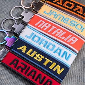May include: A collection of fabric keychains featuring embroidered names including ADAM, JAMESON, NATALIA, JORDAN, AUSTIN, and ARIANA. Each keychain has a metal ring attachment in various finishes, including silver, black, and an iridescent rainbow metal.