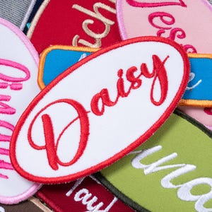 May include: An oval-shaped white fabric patch with a red embroidered border and the name Daisy stitched in red cursive script. The patch is layered among other colorful embroidered name patches in various shapes and colors including pink, blue, and green.