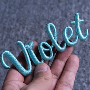 May include: A light blue embroidered patch with the word "Violet" in a cursive script. The patch is made of a soft, textured material and is held in a hand against a gray background.