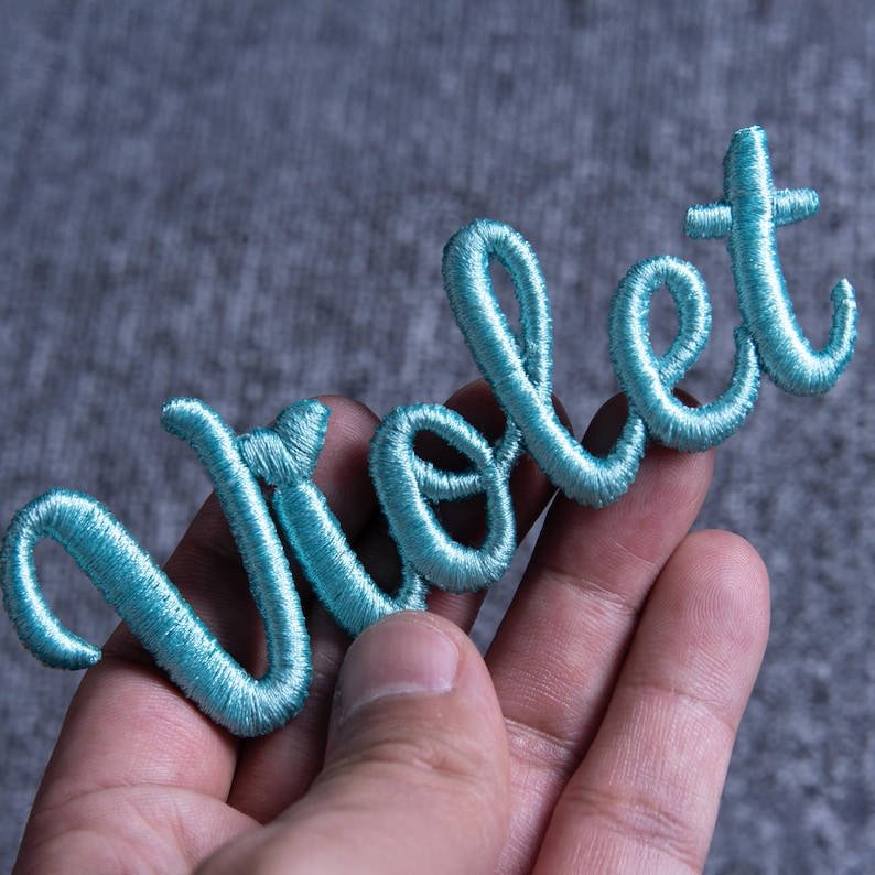 May include: A light blue embroidered patch with the word "Violet" in a cursive script. The patch is made of a soft, textured material and is held in a hand against a gray background.