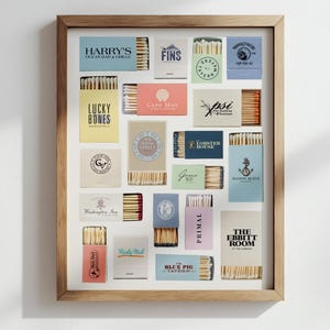 May include: A framed collection of matchboxes, each with unique designs and text. The boxes are arranged in a grid, showcasing various colors and fonts. The frame is a light brown color, and the background is white.