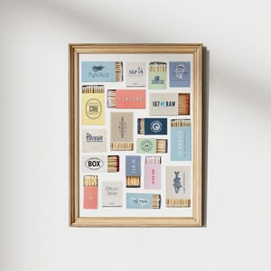 May include: A framed art print featuring a collection of matchboxes with various designs and text. The matchboxes are arranged in a grid pattern, showcasing a variety of colors and branding. The frame is a light wood color, adding a natural touch to the display.