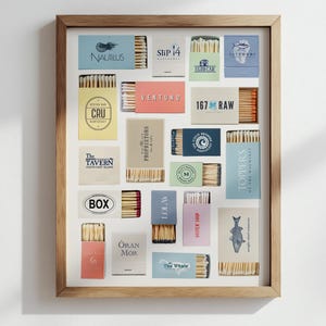 May include: A framed display featuring various matchboxes with different logos and text. The matchboxes are arranged in a grid pattern, showcasing a variety of colors and designs. The frame is made of light-colored wood.