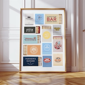May include: A framed collection of matchboxes with various designs and text. The matchboxes feature logos and text such as "Black Whale," "Hudson House Bar," "Oyster House," and "Parker's Garage." The frame is wooden, and the background is a white wall and wooden floor.