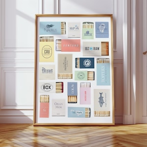 May include: A framed print featuring a collection of matchboxes with various designs and logos. The matchboxes are arranged in a grid, showcasing a range of colors and branding. The frame is a warm, natural wood tone, and the background is a white wall with a wooden floor.