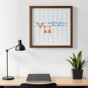 May include: A framed print featuring a watercolor illustration of a bikini top and bottom hanging on a clothesline. The background has blue and white vertical stripes, with the text "meet me down the shore" in blue script.