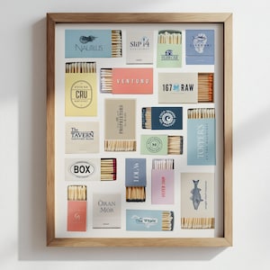 May include: A framed display featuring various matchboxes with different logos and text. The matchboxes are arranged in a grid pattern, showcasing a variety of colors and designs. The frame is made of light-colored wood.