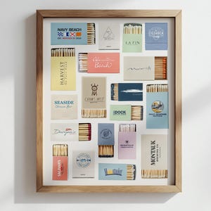 May include: A framed collection of matchboxes, each with a unique design and text. The boxes are arranged in a grid, showcasing various colors and fonts. The frame is a light brown color.