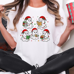 Peut inclure: T-shirt blanc avec un motif de six fantômes de dessins animés portant des chapeaux de Père Noël et lisant des livres. Les fantômes sont décorés de guirlandes lumineuses et d'étoiles. Un design festif pour la période des fêtes.